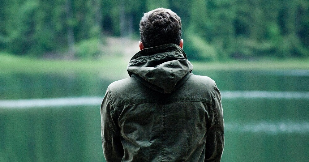 A man wearing a hooded sweater, standing by the water in the woods
