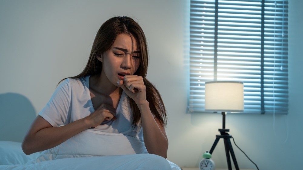 Young Asian woman sitting up in bed coughing