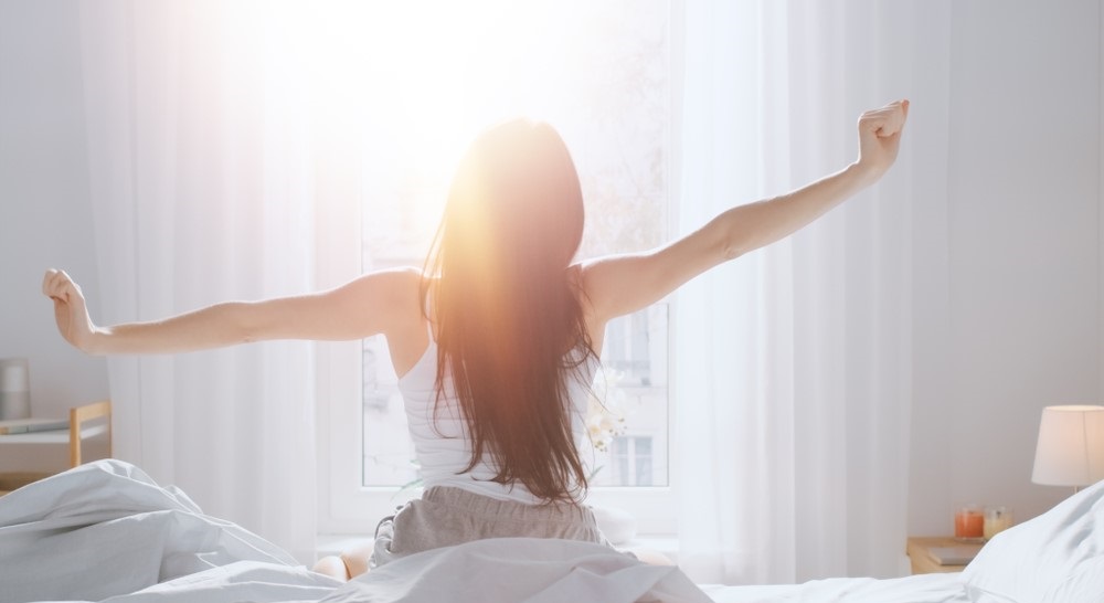 Young woman sitting up in bed stretching in morning sunlight