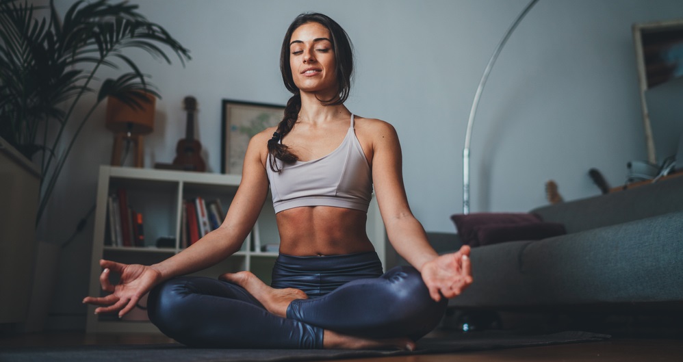 beautiful woman in sportive top and leggings practicing yoga at home sitting in lotus pose on yoga mat