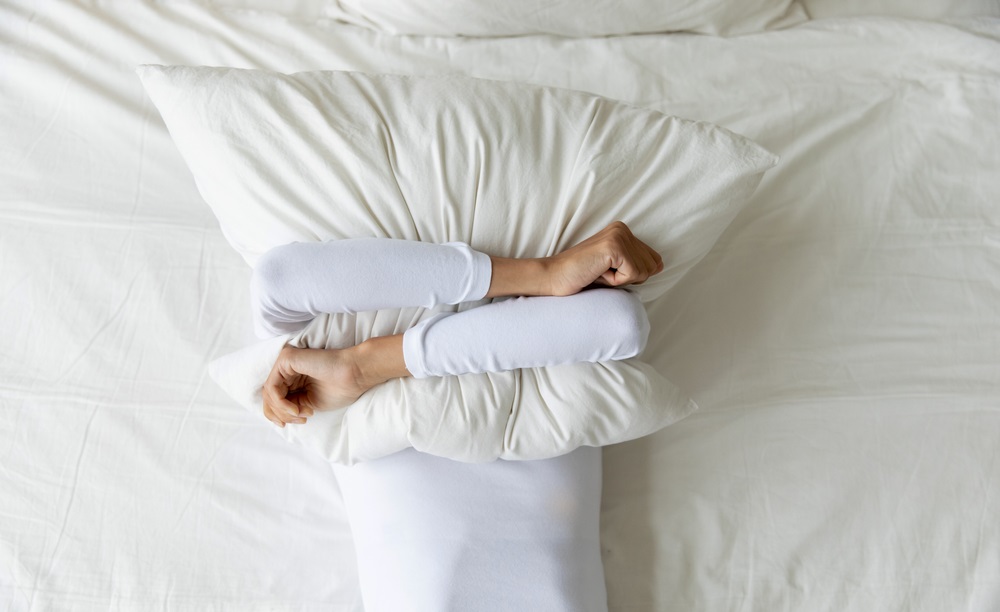 depressed woman covering face with pillow, lying on bed at home alone