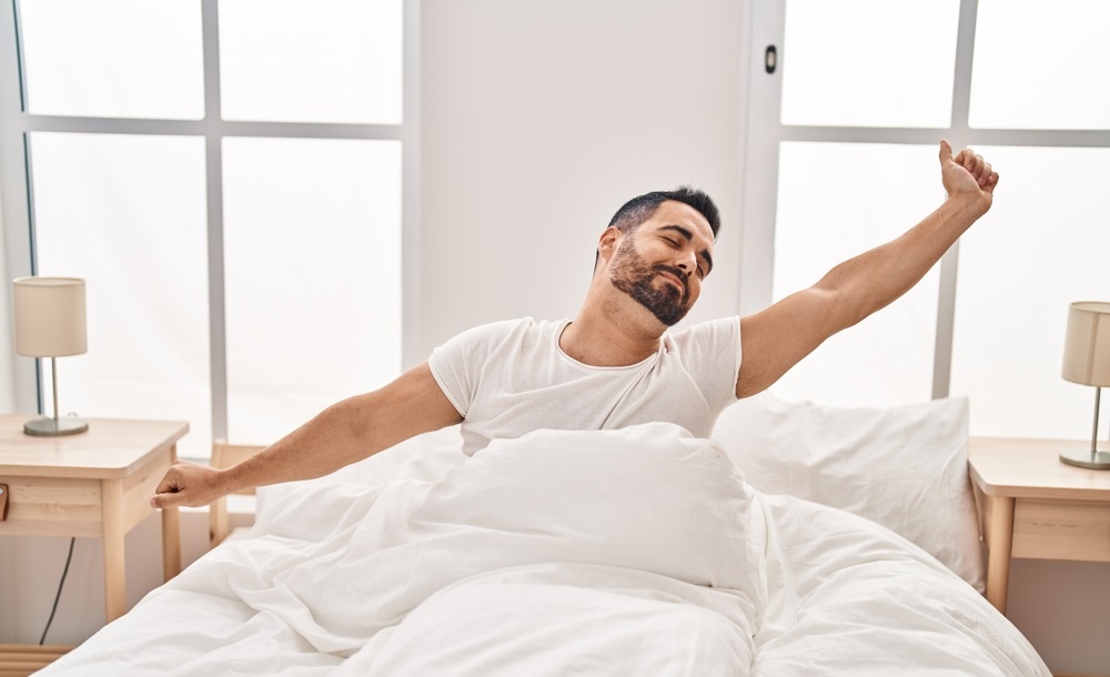 hispanic man stretching arms sitting on bed at bedroom