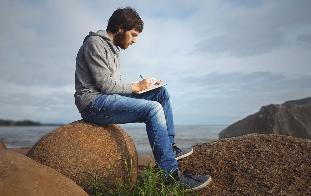 man sitting on stones near the sea and writing on notebook