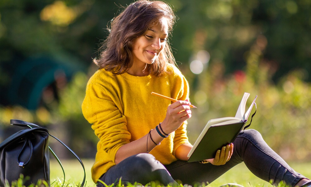 smiling young woman sitting on grass in park writing in book